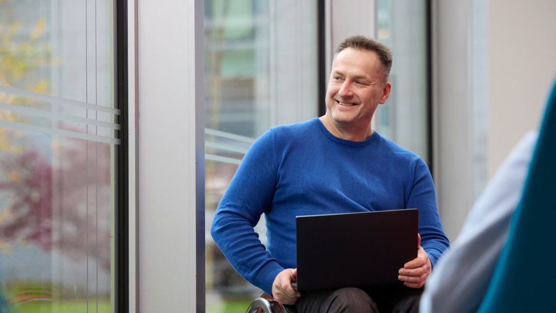 Jan Hecker, Mitarbeiter der KfW Bankengruppe, trägt einen blaune Pullover und sitzt in enem Rollstuhl. Er hat einen schwarzen Laptop auf dem Schoß und hält ihn mit den Händen rechts und links fest. Vor ihm steht ein niedriger, runder ,schwarzer Tisch. Hinter ihm befindet sich eine Fensterfront, durch die herbstlich gefärbte Grünflächen zu sehen sind. 