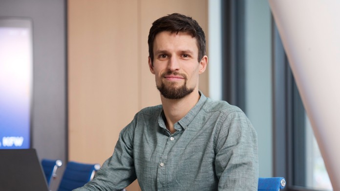 Johannes Hengst, Mitarbeiter der KFW Bankengruppe, sitzt an einem Konferenztisch auf einem blauen Stuhl. Vor ihm steht ein aufgeklappter schwarzer Laptop. Rechts und links von ihm sind freie Stühle, ebenfalls blau. Im Hintergrund ist ein großer, blauer Displaymit dem Logo der KfW Bankegruppe zum Teil zu sehen.