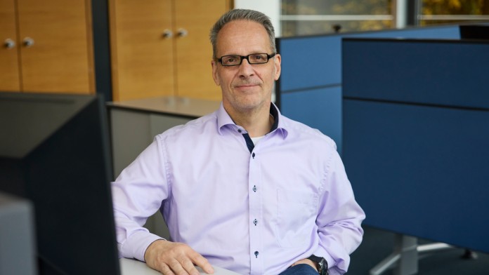 Jörg Wycisk, an employee of the KfW banking group, is sitting at a desk with a computer. He is wearing a light blue shirt, glasses, and blue jeans. One hand is in his pocket and the other rests on the table. In the background, blue partition walls and two wooden cabinets can be seen.