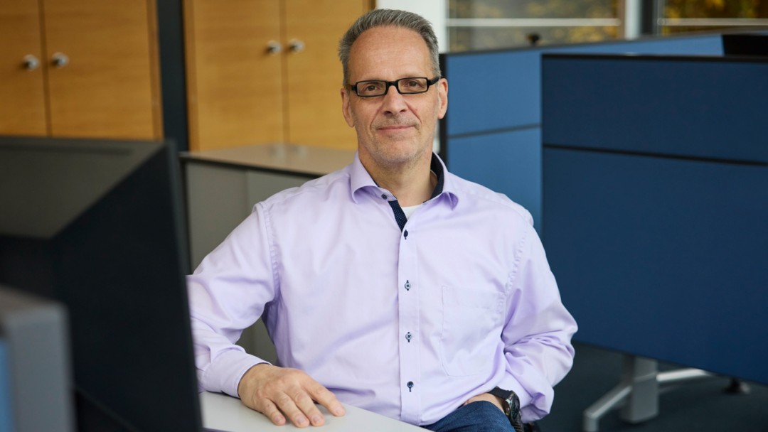 Jörg Wycisk, Mitarbeiter der KfW Bankengruppe, sitzt an einem Schreibtisch mit einem Computer. Er trägt ein hellblaues Hemd, eine Brille und eine blaue Jeanshose. Eine Hand steckt in der Hosentasche und die andere ruht auf dem Tisch. Im Hintergrund sind blaue Trennwände und zwei Holzschränke zu sehen. 