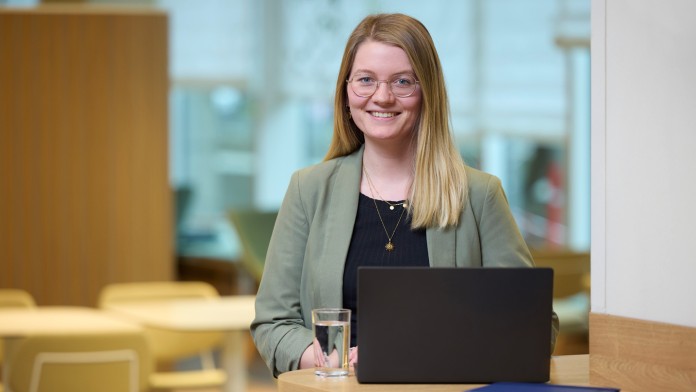 Frau Luisa Struck steht in einem hellen Raum an einem Tisch, auf welchem ein aufgeklappter Laptop, ein Glas Wasser und ein blaues Notizbuch steht. Sie hat blonde Haare, trägt eine Brille, einen olivgrünen Blazer und schaut freundlich in die Kamera.