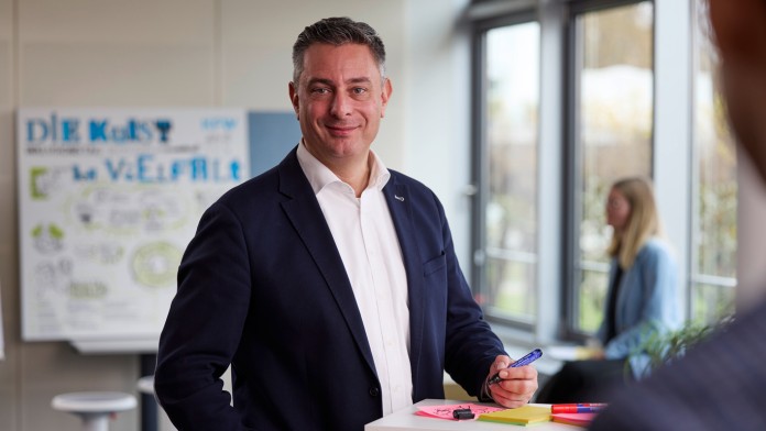 The representative for severely disabled employees at KfW, Martin Höfel, is looking friendly into the camera. He has short dark hair and is wearing a white shirt and a dark blue jacket. He is standing at a table on which a labeled sticky note is lying. In the background, a blonde-haired colleague can be seen looking at a flip chart with the heading “Diversity.”