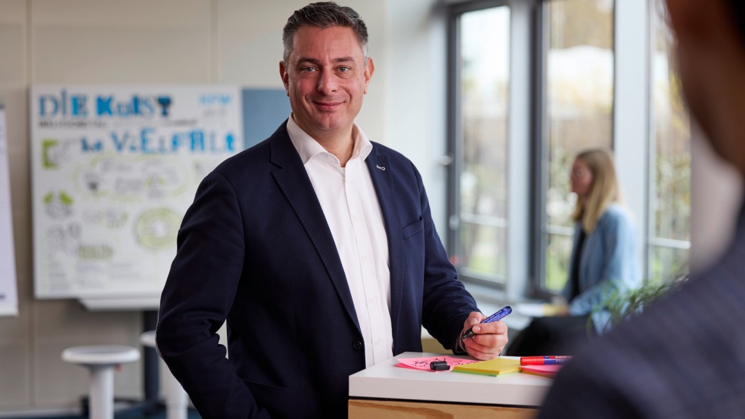 Der Schwerbehindertenvertreter der KfW, Martin Höfel, schaut freundlich in die Kamera. Er hat kurze dunkle Haare und trägt ein weißes Hemd sowie ein dunkelblaues Sakko. Er steht an einem Tisch, auf dem eine beschriftete Haftnotiz liegt. Im Hintergrund ist eine Kollegin mit blonden Haaren zu sehen, die auf ein Flipchart mit der Überschrift 'Vielfalt' blickt.