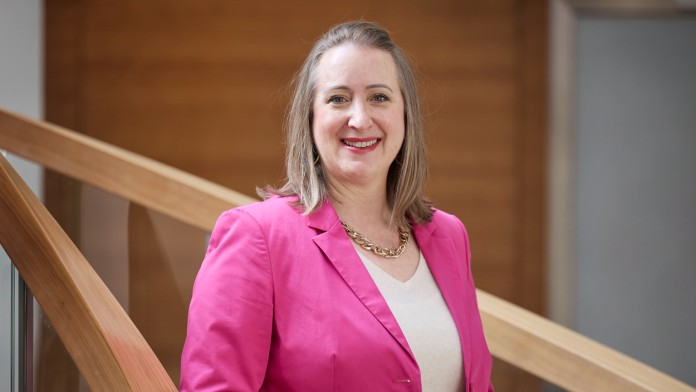 Nina Poppenhäger, an employee of the KfW banking group, is standing smiling on a staircase with her hands in her pockets. She is wearing a pink blazer over a light, thin V-neck sweater. She has shoulder-length hair and is wearing a necklace.
