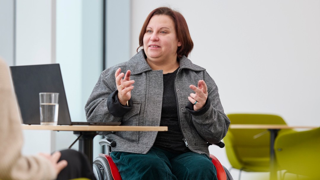 Nora Schratz, Mitarbeiterin der KfW Bankengruppe, sitzt in einem hellen Raum in einem Rollstuhl. Vor ihr ist ein runder Tisch, ein aufgeklappter Laptop und ein Glas Wasser. Es sitz ihr gegenüber eine blonde Frau auf einem grünen Stuhl. Nora Schratz hat rötliche, schulterlange Haare, eine graue Jacke und eine petrolfarbene Hose an. Im Hintergrund ist ein weiterer runder Tisch mit grünen Stühle zu sehen. 