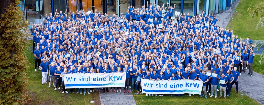 Auf dem Bild ist eine große Gruppe von Mitarbeitenden der KfW vor einem Gebäude zu sehen. Alle Personen tragen blaue Oberteile und strecken die Hände nach oben. In der ersten Reihe sind zwei Banner zu erkennen, auf denen „Wir sind eine KfW“ steht.