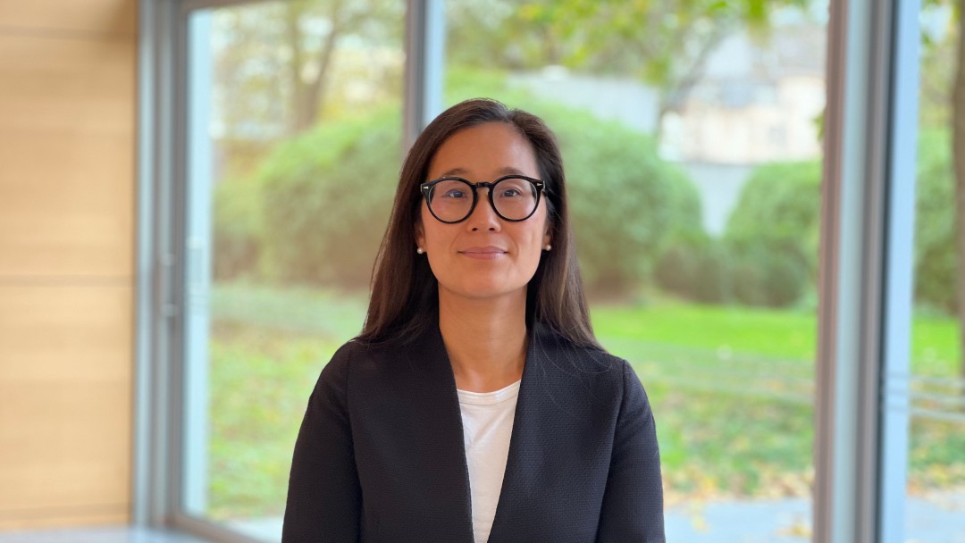 Hae-Young Koo, Mitarbeiterin des Inhouse Consultings der KfW, steht in einem Foyer. Sie trägt eine schwarze runde Brille, ein helles Oberteil mit dunklem Blazer.