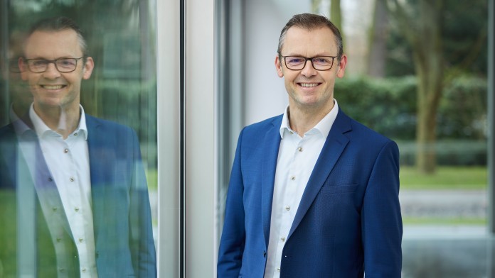 Carsten Heineke im blauen Anzug und weißem Hemd steht lächelnd mit Brille vor einer Fensterfront mit seinem Spiegelbild. 