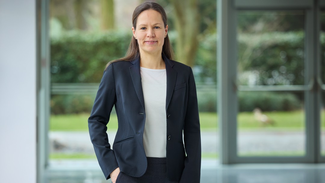 Gesa Lüdemann in einem dunklen Anzug und hellem T-Shirt, steht mit einer Hand in der Hosentasche vor einer hellen bodentiefen Fensterfront mit Gartenblick. 
