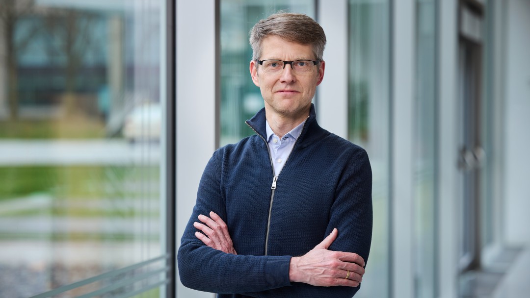 Jürgen Hinxlage mit verschränkten Armen vor einer Fensterfront, trägt ein hellblaues Hemd, einen dunkelblaune Sweater und eine Brille. 