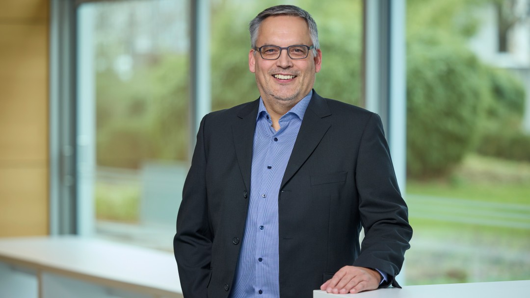 Matthias Rauch steht vor einem Hochtisch, auf dem seine linke Hand ruht. Er trägt ein blaues Hemd , einen dunklen Blazer, eine Brille und lächelt freundlich. 