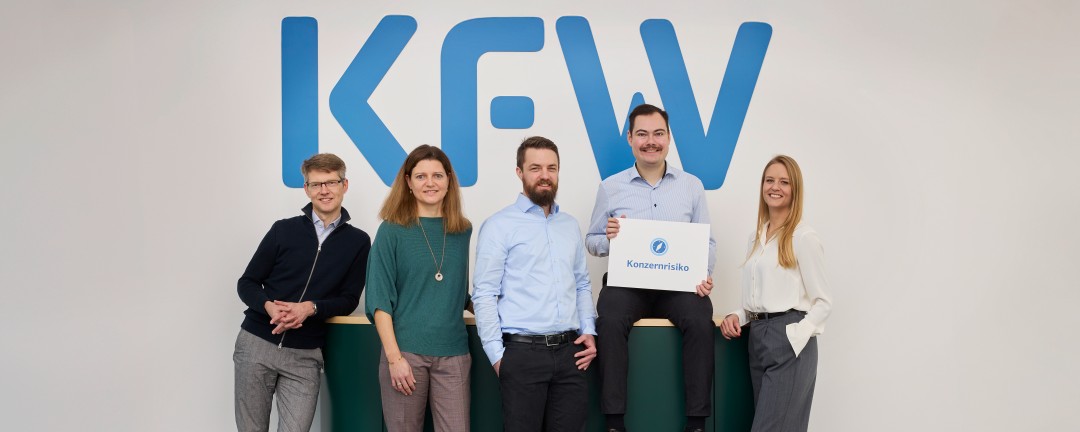 Fünf Mitarbeitende der KfW, davon zwei Frau und drei Männer, stehen gemeinsam vor einem Sideboard. Einer der Männer hält ein Schild mit der Aufschrift „Konzernrisiko“. Im Hintergrund ist eine weiße Wand mit großem blauem KfW-Logo zu sehen.