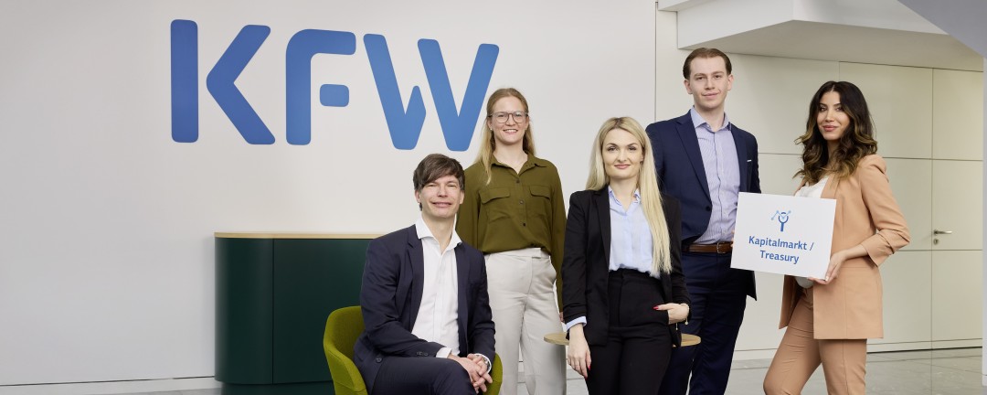 Fünf Mitarbeitende der KfW, davon drei Frauen und zwei Männer, posieren vor einer weißen Wand mit blauem KfW-Logo. Einer der Männer sitzt, die übrigen stehen. Eine der Frauen hält ein Schild mit der Aufschrift „Kapitalmarkt und Treasury“. 