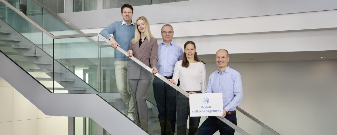 Fünf Personen, davon drei Männer und zwei Frauen, stehen im Wechsel auf einer Treppe. Der Mann auf der untersten Stufe hält ein weißes Schild mit blauer Aufschrift „Modellrisikomanagement“.