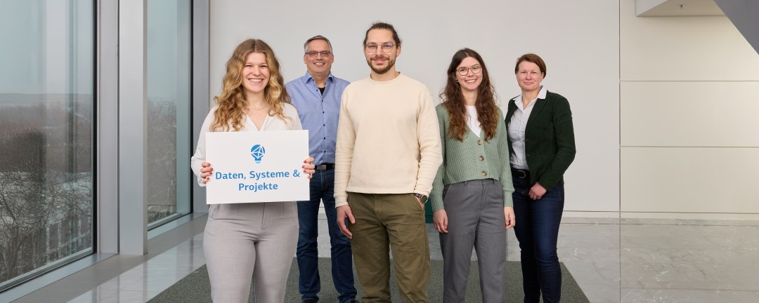Fünf Teammitglieder, davon drei Frauen und zwei Männer, stehen auf einem grauen Teppich in einem großen, hellen Raum. Die Frau in der vordersten Reihe hält ein weißes Schild mit blauer Aufschrift „Datensysteme und Projekte“. 