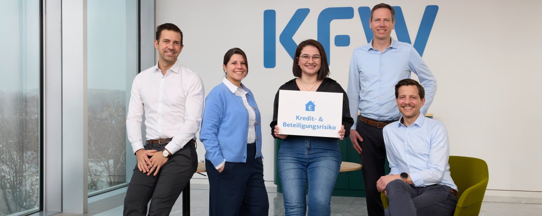 Fünf Personen, davon zwei Frauen und drei Männer, bilden ein Team, das sich vor einer weißen Wand mit blauem KfW-Logo positioniert hat. Ein Mann und eine Frau lehnen an einem Tisch, ein weiterer Mann sitzt. Eine Frau in der Mitte hält ein weißes Schild mit der blauen Aufschrift „Kredit- und Beteiligungsrisiko“. 