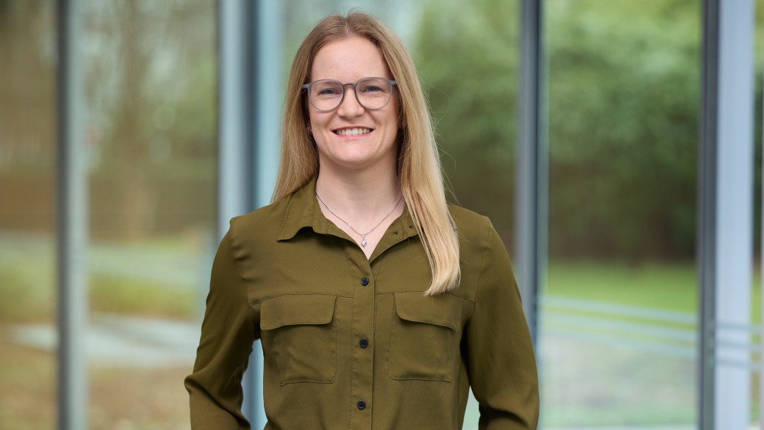Vanessa Hille steht mit beiden Händen in der Hosentasche vor einer Fensterfront. Sie hat lange blonde Haare und trägt eine Brille, sowie eine helle Hose und ein Hemd in der Farbe Khaki. 