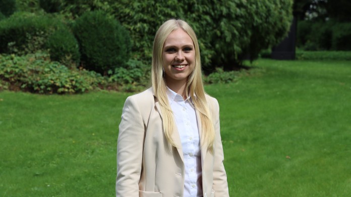 Portraitbild von Chiara Zsembera, Mitarbeiterin im Rechnungswesen der KfW, sie trägt ein helles Oberteil sowie einen hellbeigen Blazer und steht in einem Innenhof der KfW.