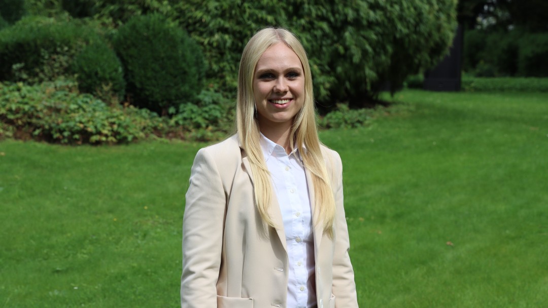 Portraitbild von Chiara Zsembera, Mitarbeiterin im Rechnungswesen der KfW, sie trägt ein helles Oberteil sowie einen hellbeigen Blazer und steht in einem Innenhof der KfW.