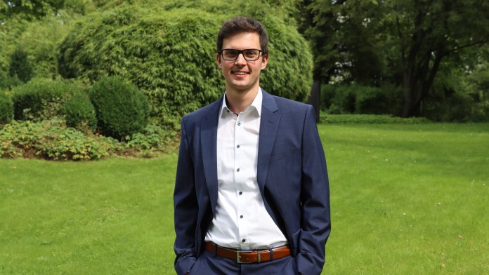 Portraitbild Eric Reiners, Trainee im Rechnungswesen der KfW, steht in einem Innenhof der KfW. Er trägt eine Brille und hat ein weißes Hemd mit dunkelblauem Anzug und braunem Gürtel an. Beide Hände sind locker in die Hosentasche gesteckt.