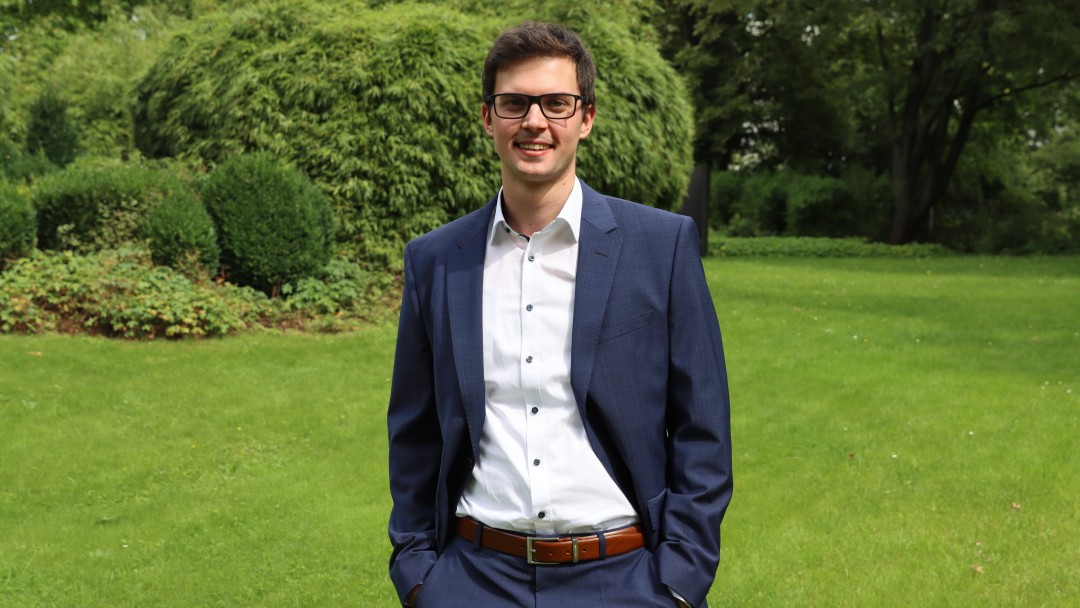 Portraitbild Eric Reiners, Trainee im Rechnungswesen der KfW, steht in einem Innenhof der KfW. Er trägt eine Brille und hat ein weißes Hemd mit dunkelblauem Anzug und braunem Gürtel an. Beide Hände sind locker in die Hosentasche gesteckt.