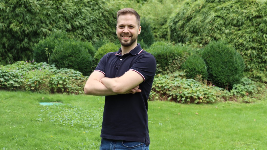 Portraitaufnahme von Niklas Kunkel, Mitarbeiter im Rechnungswesen der KfW, er steht in einem Innenhof der KfW, trägt ein dunkelblaues Poloshirt und Jeans, die Arme sind gekreuzt.