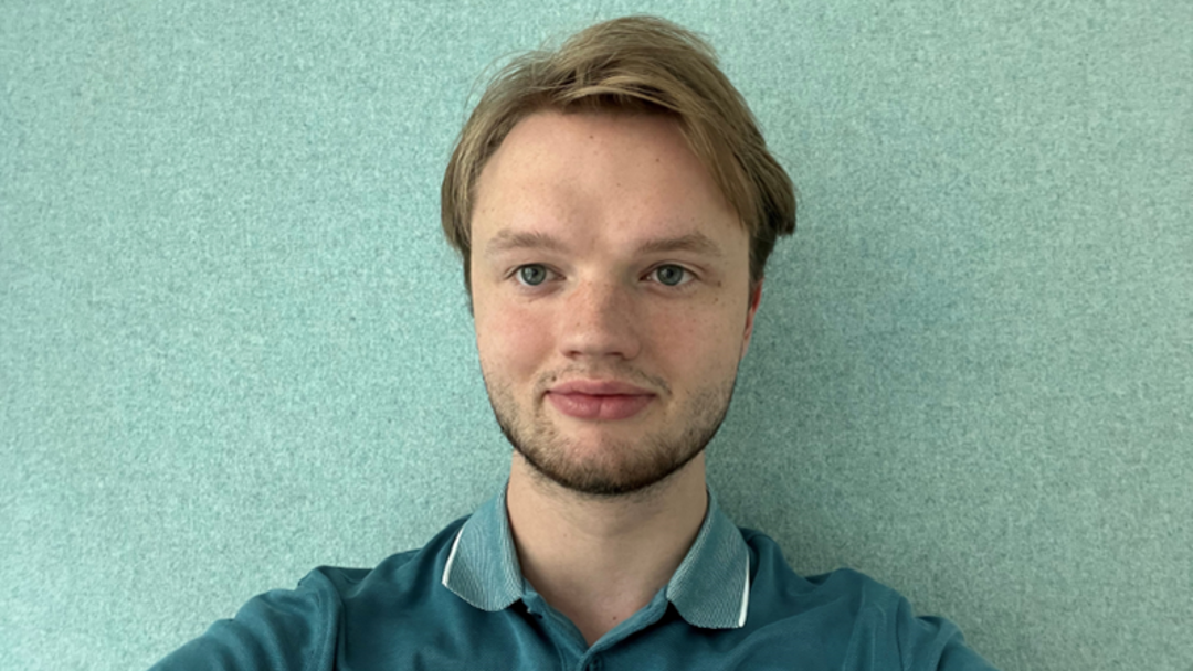 Der Mitarbetiter Johann Koch steht vor einer hellgrünen Wand, hat blonde, kurze Haare und trägt ein Grünes Polo-Tshirt an.