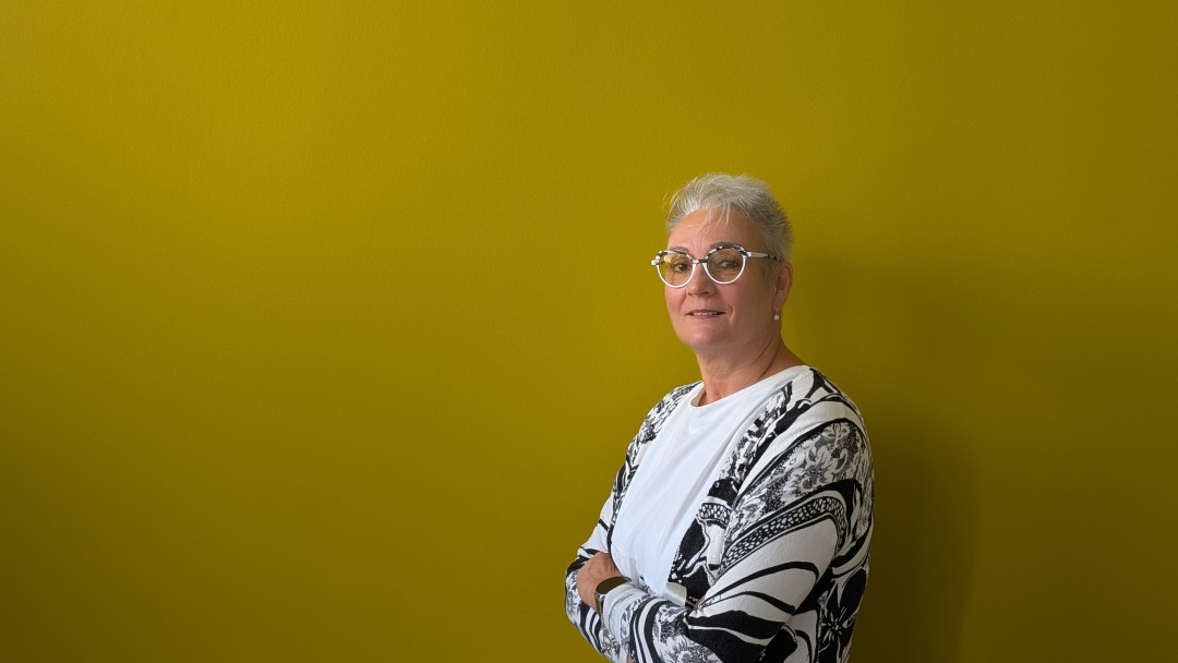 Auf dem Bild ist eine Frau mit weißem Oberteil und Brille zu sehen, die vor einem ockerfarbenen Hintergrund steht. Sie hat die Arme verschränkt und lächelt. 