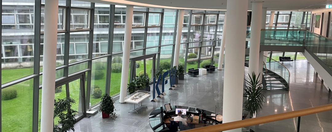 Empfangsbereich der KfW-Niederlassung in Bonn in einem großen verglasten Foyer mit grauen Fliesen und weißen Balken. Eine Person sitzt am Empfang. Im Hintergrund sieht man eine Grünfläche. 