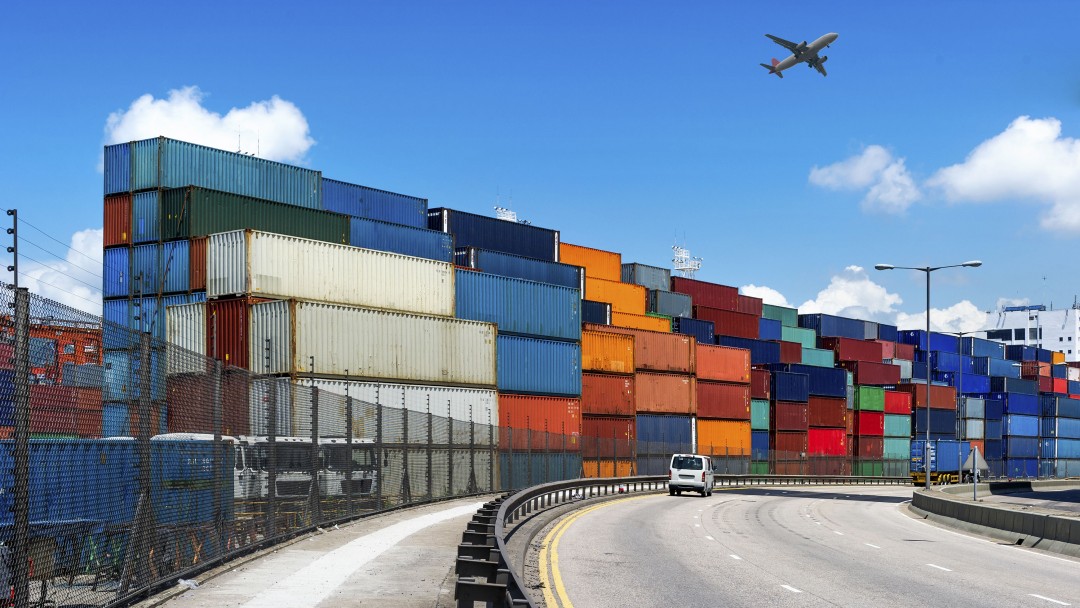 Container aufgestapelt, im Vordergrund eine Straße und in der Luft ein Flugzeug