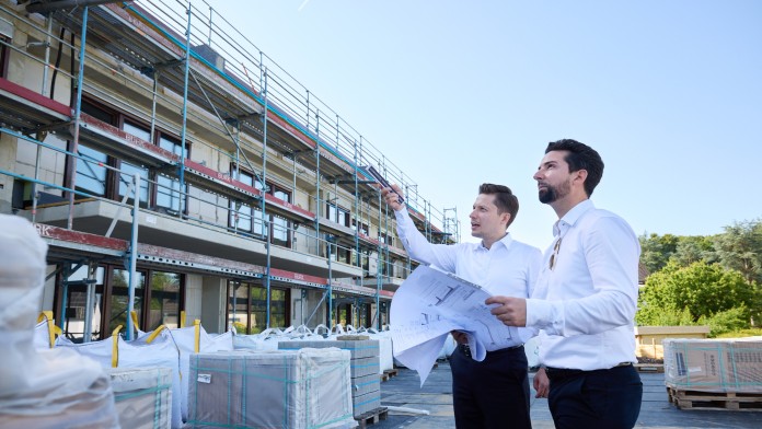 Zwei Männer in weißem Hemd stehen vor einer Baustelle und betrachten Baupläne, einer zeigt mit dem Finger auf das Gebäude.