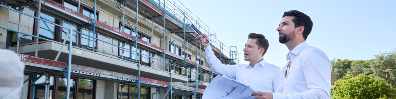 Zwei Männer in weißem Hemd stehen vor einer Baustelle und betrachten Baupläne, einer zeigt mit dem Finger auf das Gebäude.