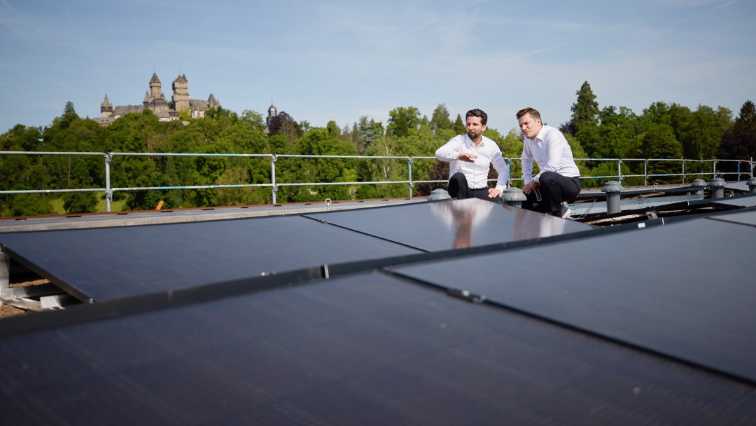 Zwei Männer in weißen Hemden sitzen auf einem Dach mit Solarpaneelen, im Hintergrund ein Schloss und Bäume unter blauem Himmel.