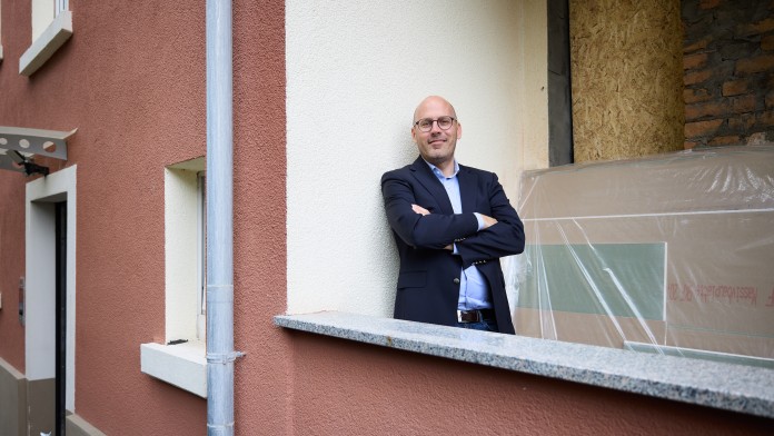 Ein Mann mit Brille und Anzug lehnt mit verschränkten Armen an einem braunen Balkon vor einem Haus mit weißem Fensterrahmen und einer mit Folie abgedeckten Öffnung.