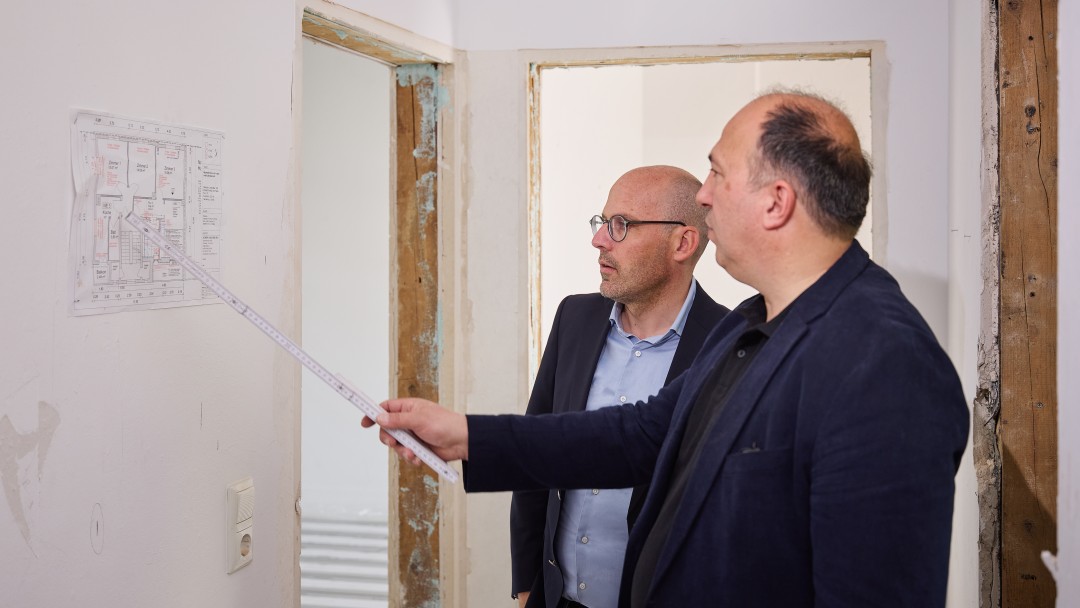 Zwei Männer in Anzügen betrachten einen Bauplan an der Wand, einer zeigt mit einem Maßstab darauf