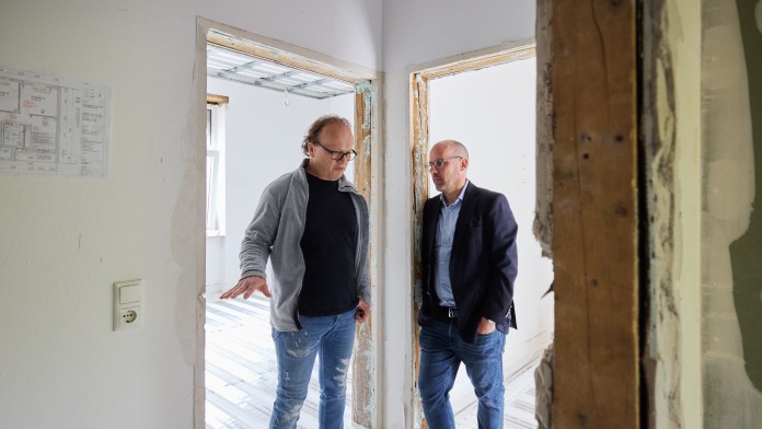 Zwei Männer in legerer und formeller Kleidung stehen in einem unfertigen Raum mit sichtbaren Trockenbauwänden und Bodenisolierung und unterhalten sich