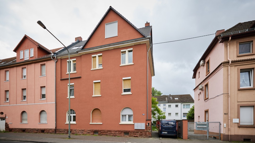 Straßenseite mit zwei mehrstöckigen Wohnhäusern und einem geparkten blauen Kleintransporter auf einem Hof zwischen den Gebäuden. Das rote Haus in der Mitte zeigt das HAus was sich in der geförderten Sanierung befindet.