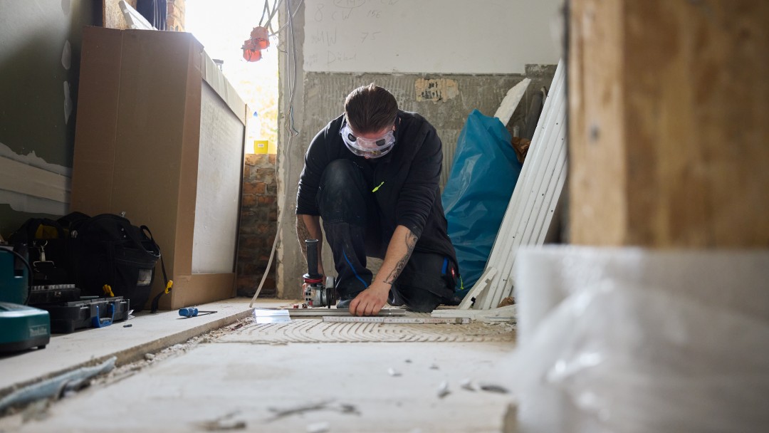 Handwerker mit Schutzbrille kniet auf Boden und arbeitet mit Elektrowerkzeug an Fliesen in einem Raum im Rohbau