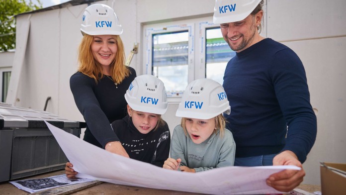 Zwei Erwachsene und zwei Kinder mit KfW-Schutzhelmen betrachten gemeinsam einen Bauplan auf einer Baustelle