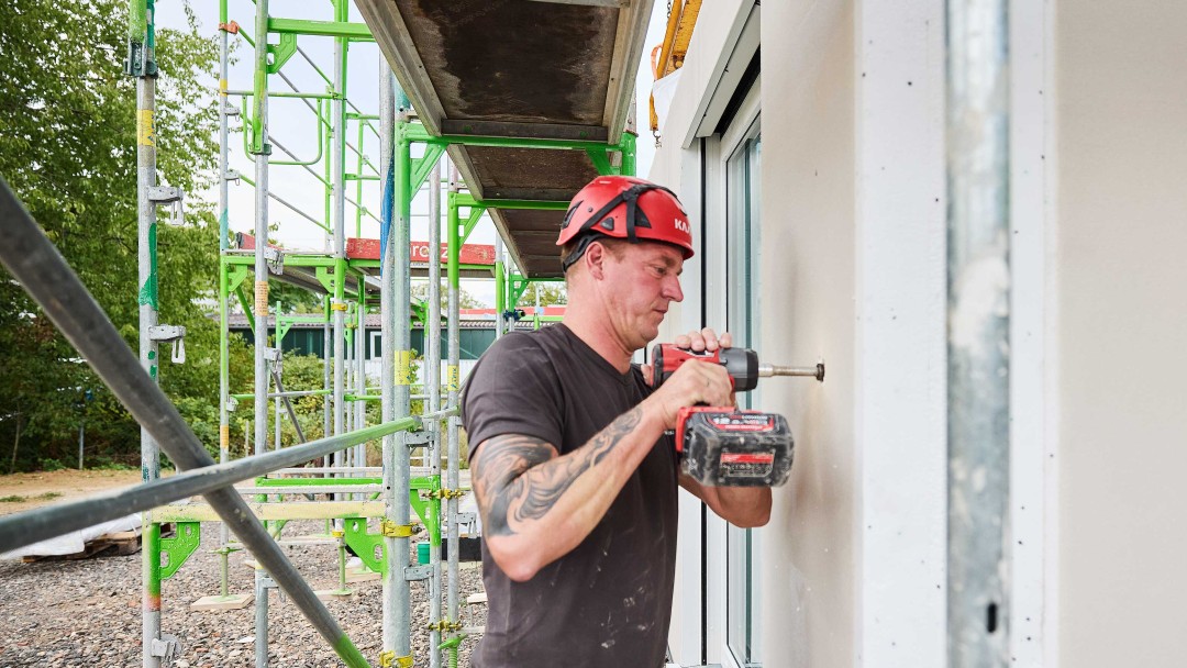 Bauarbeiter mit rotem Helm bohrt in eine weiße Wand eines Gebäudes