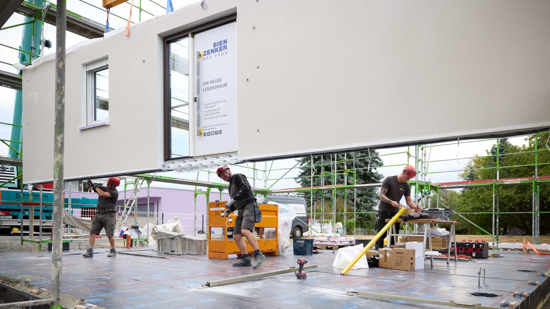 Drei Bauarbeiter mit Schutzhelmen montieren eine große vorgefertigte Wand auf einer Baustelle mit Gerüst und Kran im Hintergrund