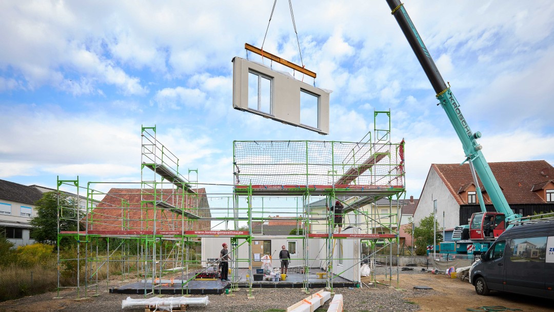 Baustelle mit Gerüst und Kran, der ein vorgefertigtes Wandsegment mit zwei Fenstern hebt, mehrere Personen stehen auf dem Boden