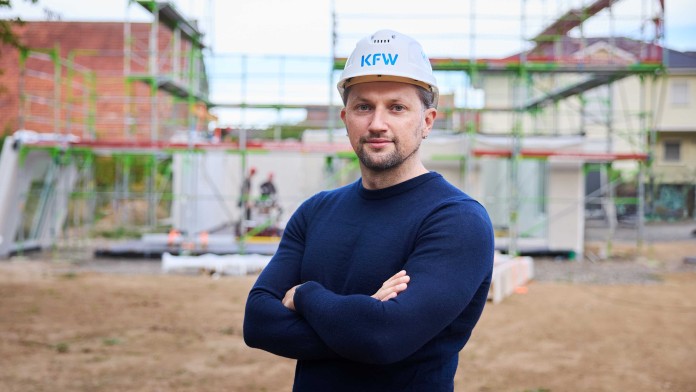 Mann mit verschränkten Armen und weißem Helm mit Aufschrift 'KfW' vor einer Baustelle mit Gerüst