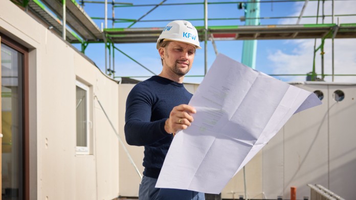 Mann mit weißem KfW-Schutzhelm liest Bauplan auf einer Baustelle mit Gerüst und blauen Himmel im Hintergrund
