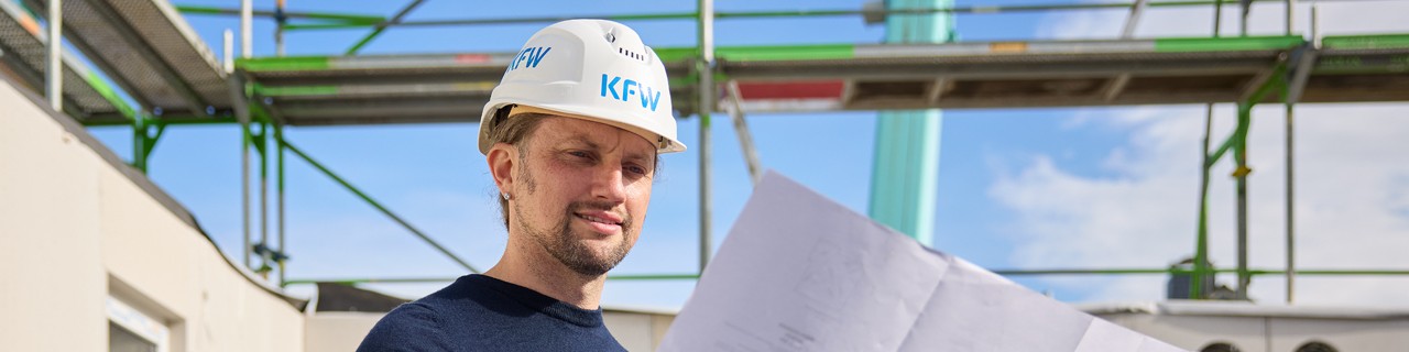 Mann mit weißem KfW-Schutzhelm liest Bauplan auf einer Baustelle mit Gerüst und blauen Himmel im Hintergrund