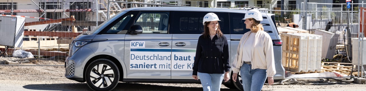 Zwei KfW Mitarbeiterinnen laufen vor einem parkenden VW Bus und einer Baustelle