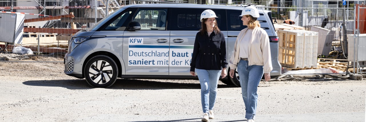 Zwei KfW Mitarbeiterinnen laufen vor einem parkenden VW Bus und einer Baustelle