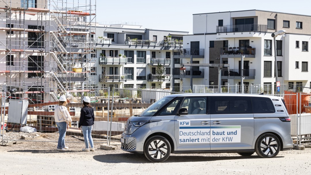 Zwei KfW Mitarbeiterinnen laufen vor einem parkenden VW Bus und einer Baustelle