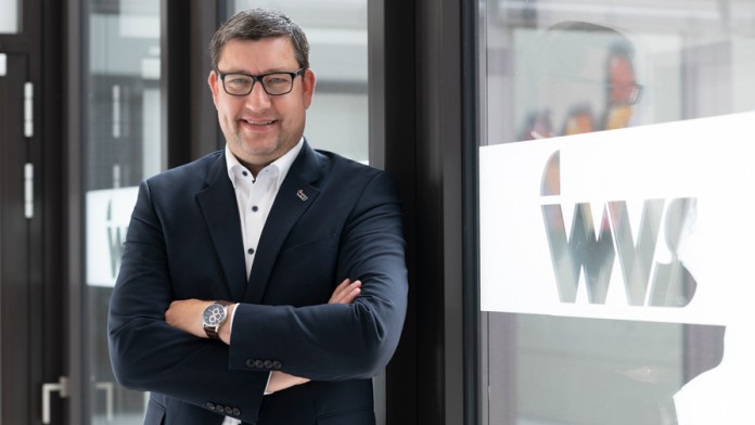 Mann mit Brille und dunklem Sakko steht mit verschränkten Armen vor einer Glaswand mit dem Schriftzug 'WVS'