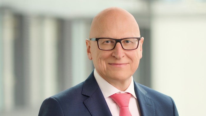 Porträt eines lächelnden Mannes mit Brille, Glatze, dunkelblauem Anzug und roter Krawatte vor unscharfem Bürogebäude